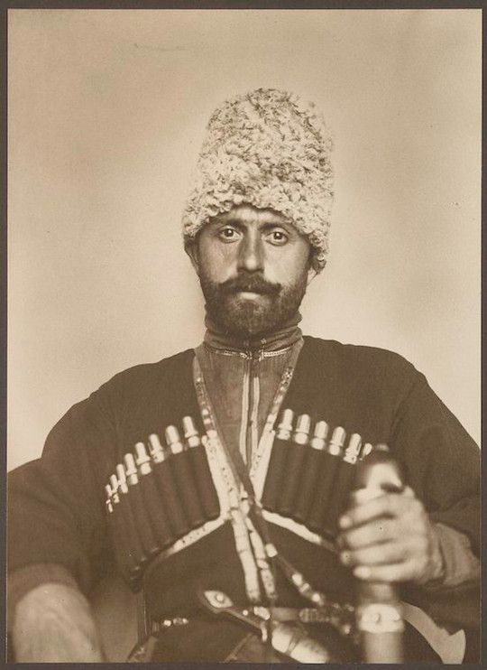 Sepia-toned portrait of a man in traditional clothing, wearing a fur hat and a cartridge belt, holding a riding crop.