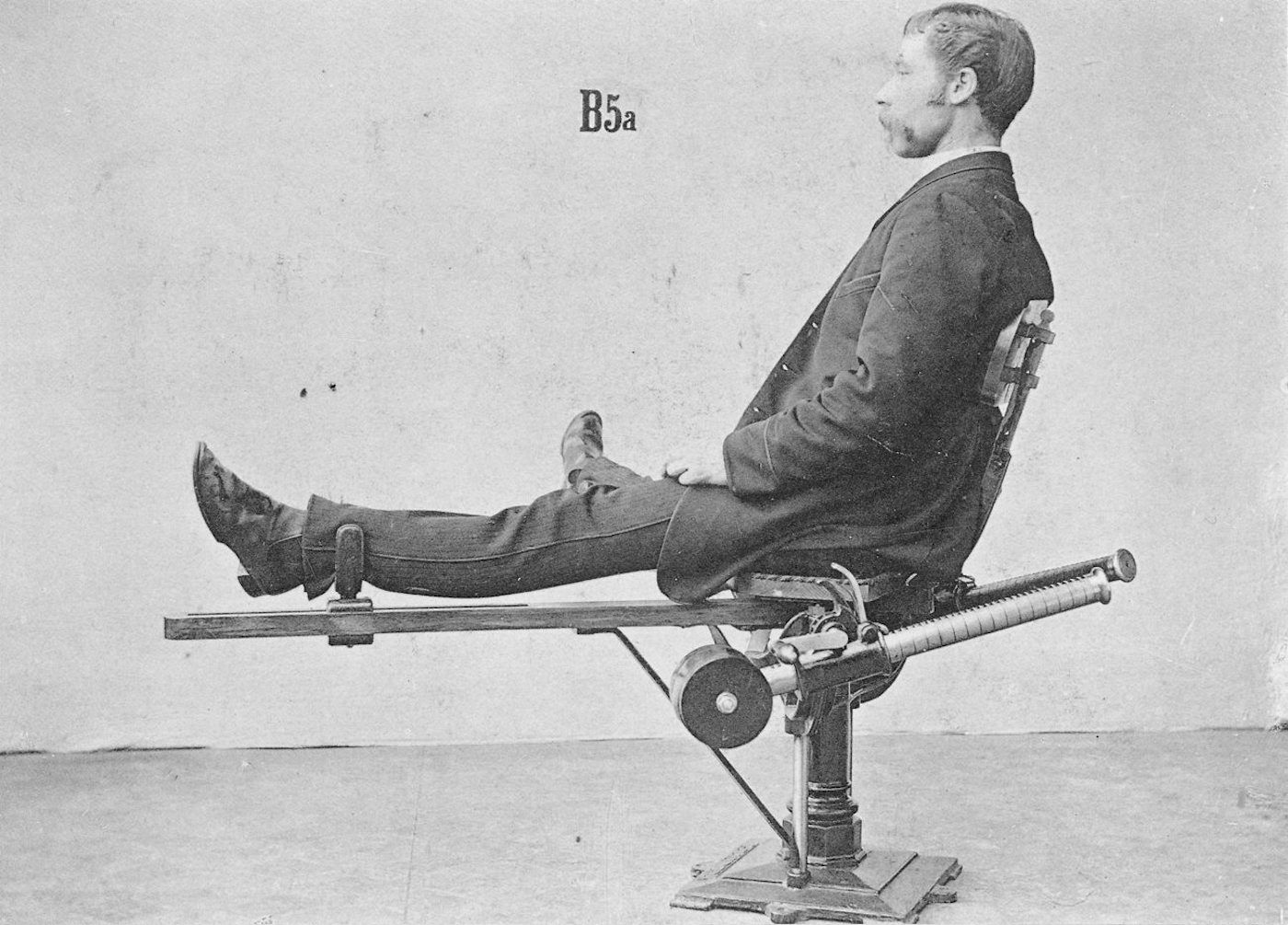 A black and white photograph of a man seated in a complex mechanical device, possibly for medical or therapeutic purposes.