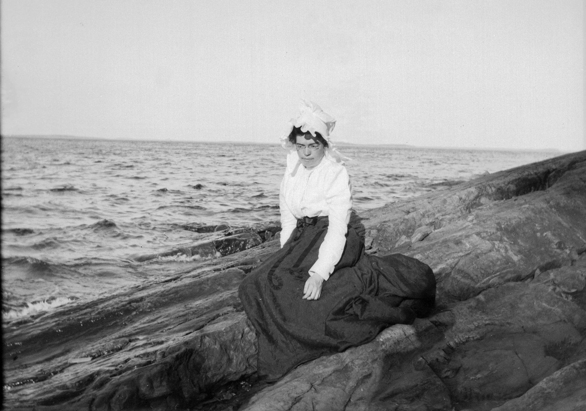 A black and white photograph of a woman sitting on a rocky outcrop overlooking the sea.
