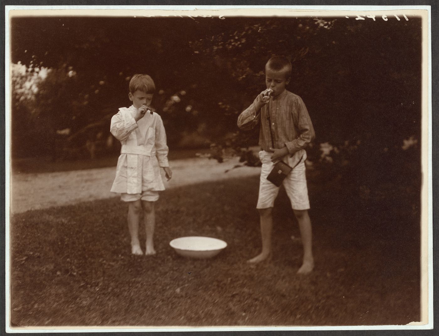 Two young boys barefoot on a lawn, drinking from a bowl.