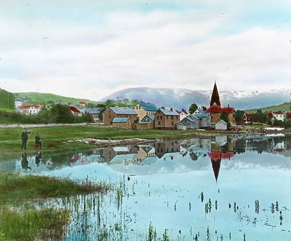 Vintage photograph of a town beside a lake with mountains in the background.