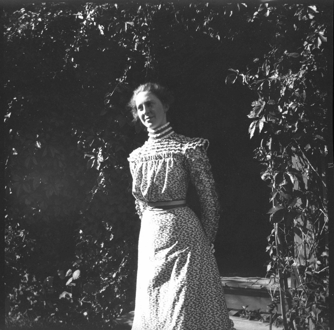 A black and white photograph of a woman in a long dress standing in front of a leafy archway.