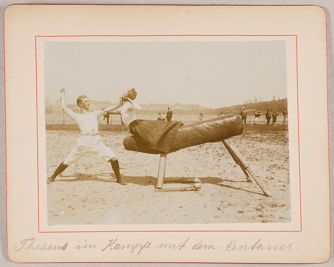 Vintage photograph of a man wrestling with a large animal, possibly a seal or sea lion, on a grassy field, with a crowd of spectators in the background.