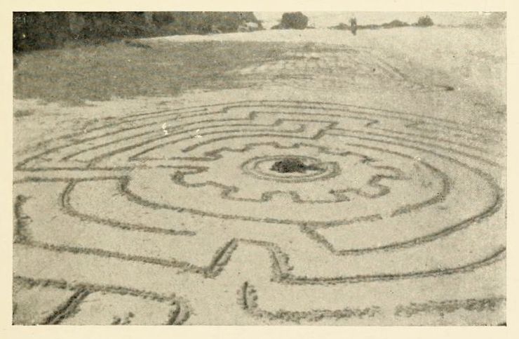 A black and white photograph of a circular maze in a field.