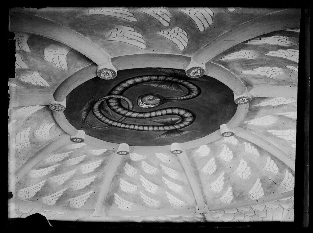 A black and white photograph of a ceiling painting featuring a dark oval with a coiled serpent design, surrounded by arched wings and leaf-like patterns.