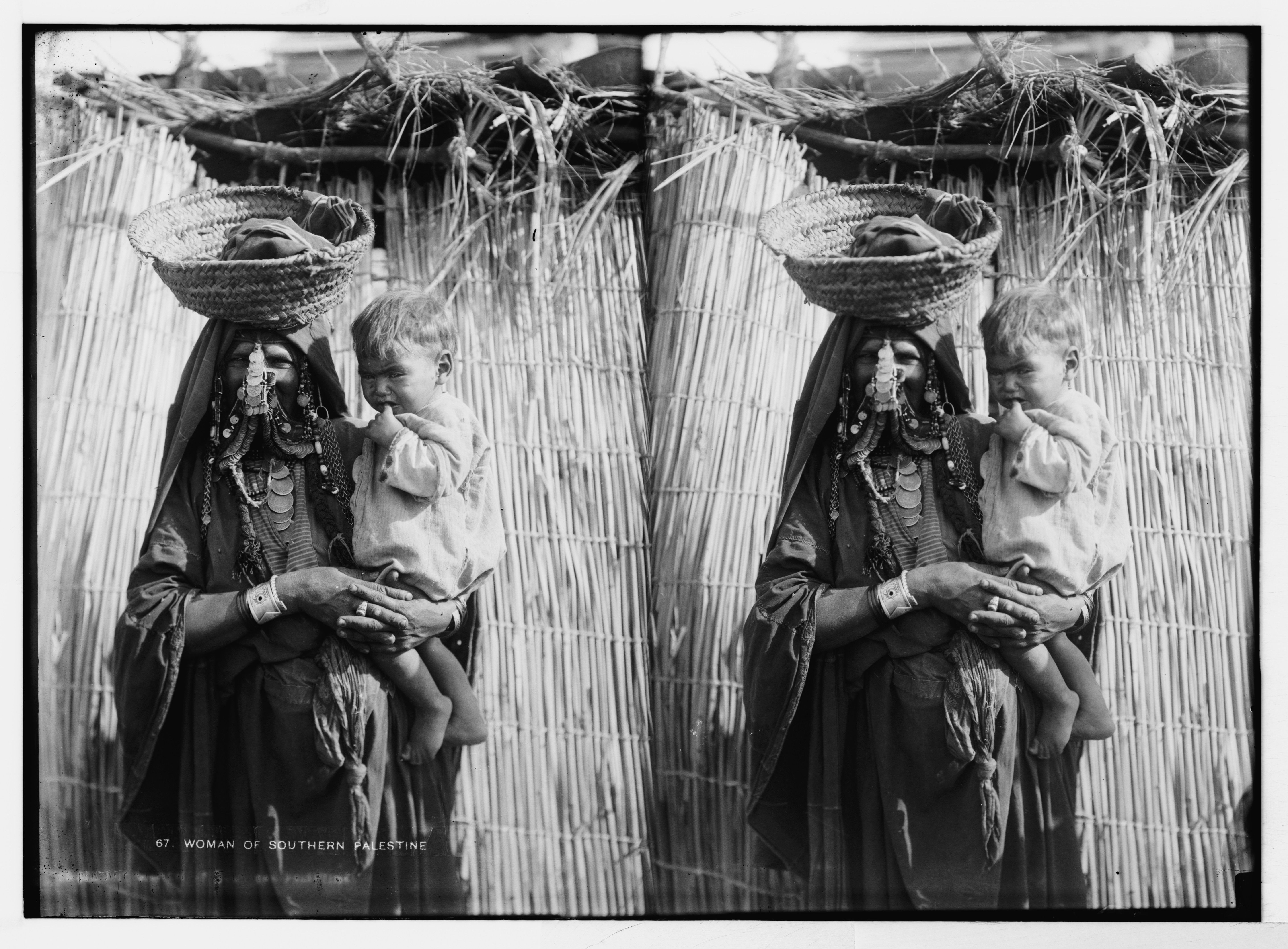 Stereoscopic photograph of a woman from Southern Palestine holding a child, dressed in traditional clothing and adorned with jewelry, against a backdrop of woven grass or straw.