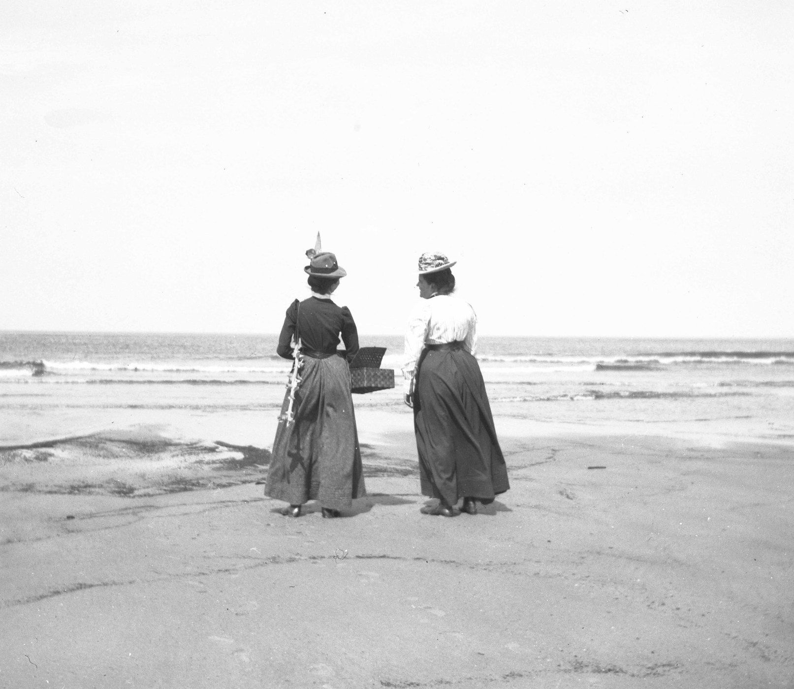 Two women in long dresses and hats stand on a beach, facing the sea, with one holding a basket.