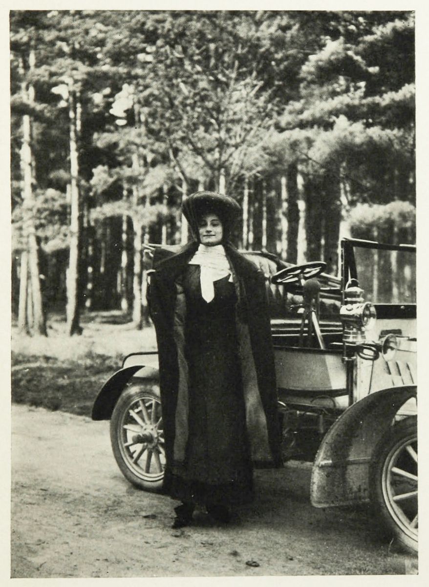 A vintage black and white photograph of a woman standing next to an early automobile in a wooded area.