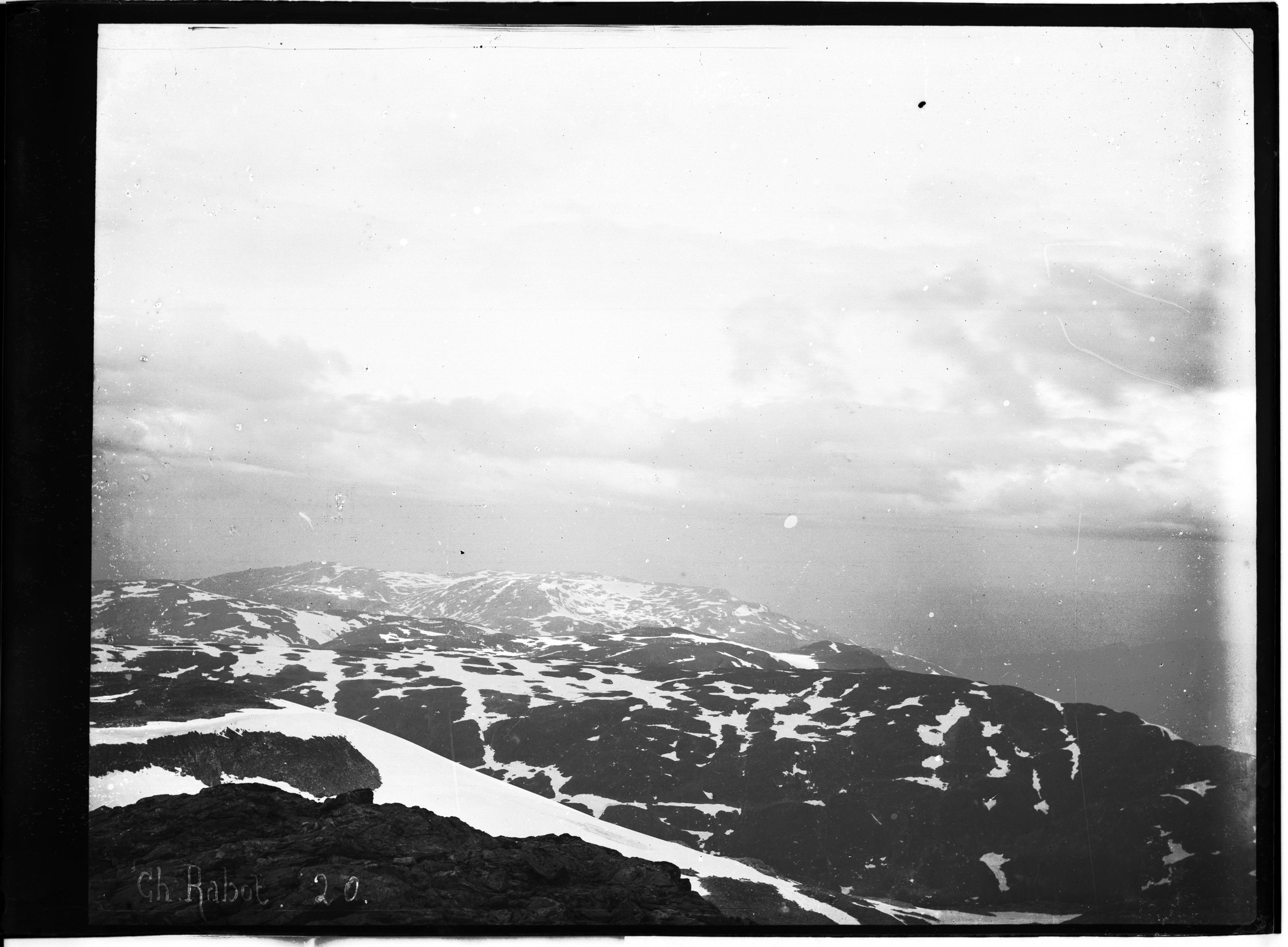 A black and white photograph of a snow-covered mountain range with a long, narrow body of water running through it. The sky is overcast with clouds.