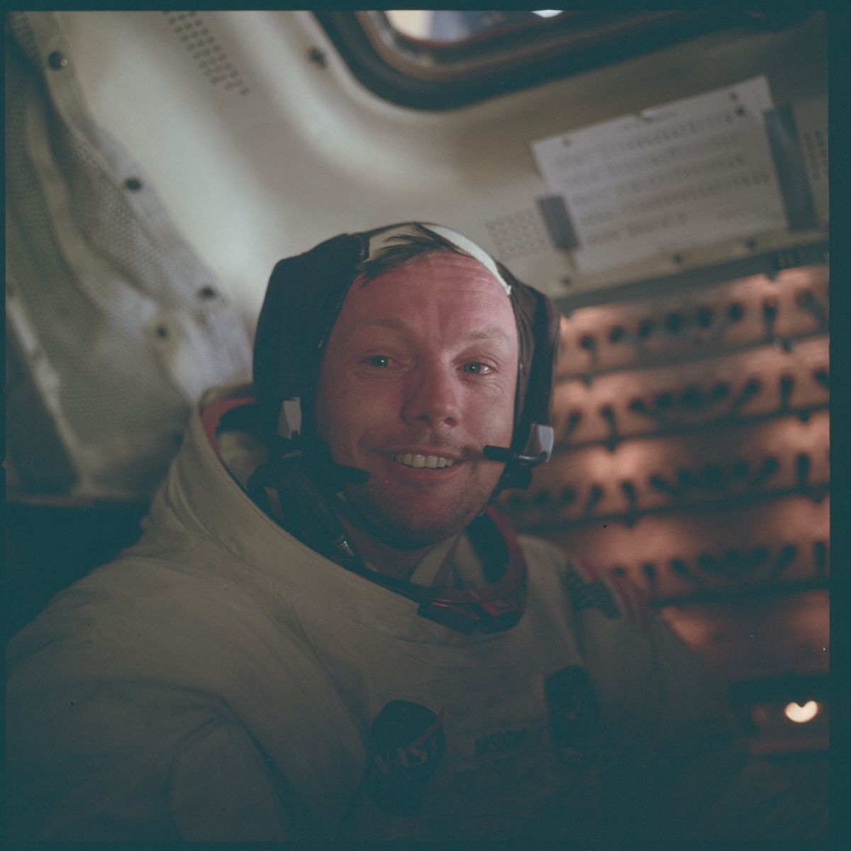 Close-up portrait of Neil Armstrong inside a spacecraft, wearing a space suit and headset, with a complex control panel in the background.