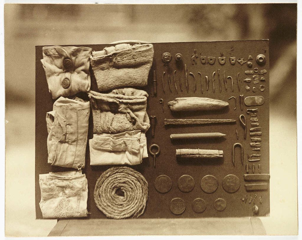 A sepia-toned photograph of a collection of old textiles and artifacts laid out on a dark, rectangular surface.