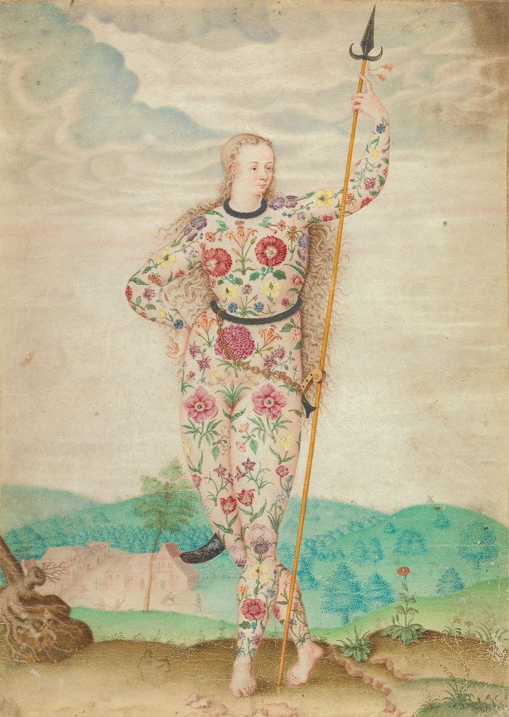 A full-body portrait of a person in a floral suit holding a spear in a landscape.