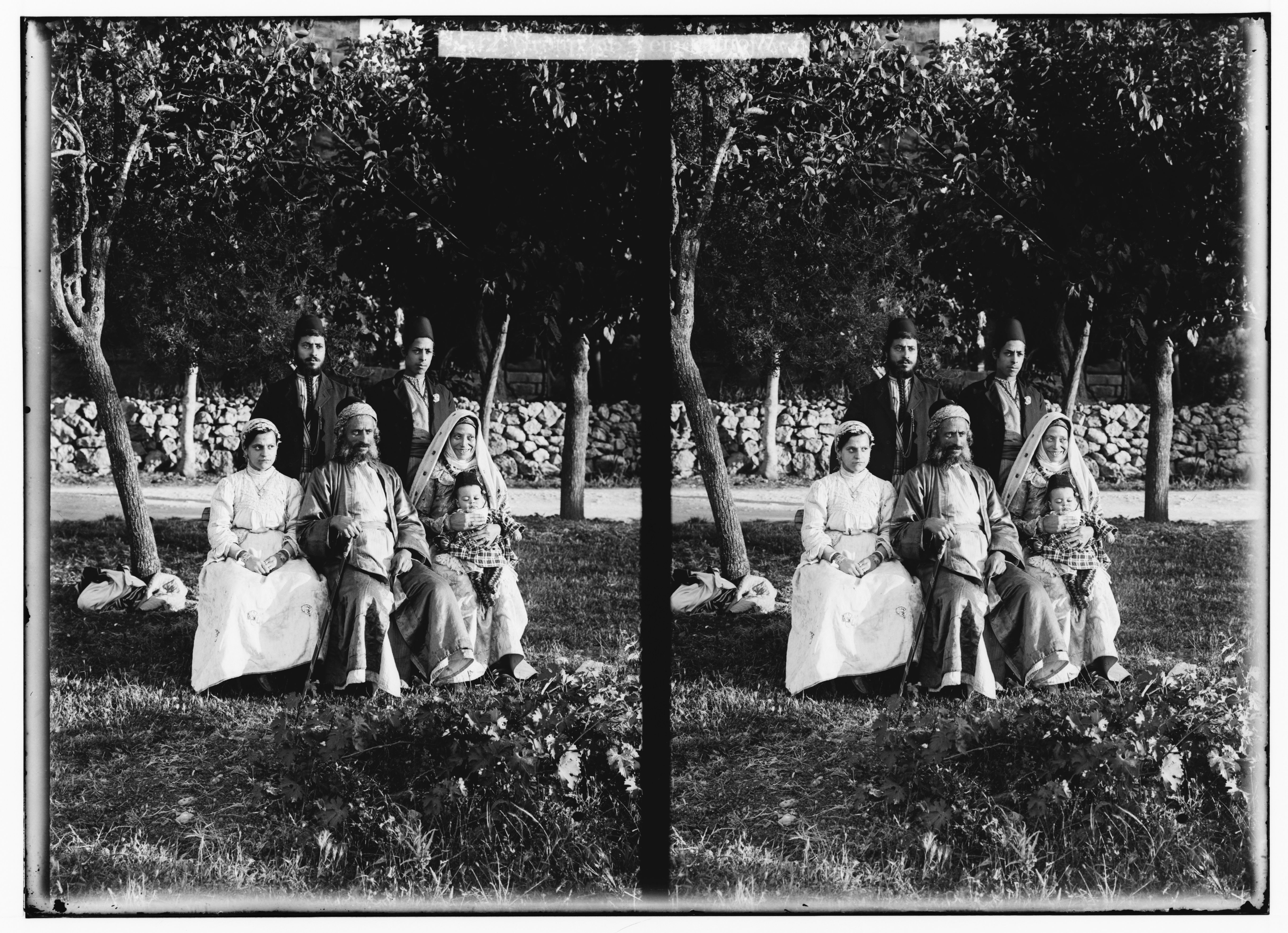 Stereoscopic photograph of a group of people posing outdoors in front of a stone wall.