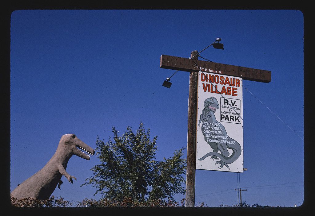 Vintage roadside attraction, Dinosaur Village, with a dinosaur statue and a sign advertising R.V. park, groceries, and other supplies.