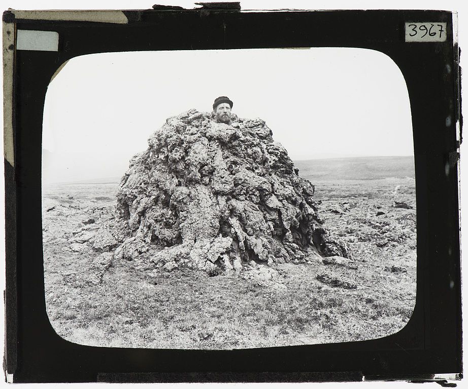 A black and white photograph of a person almost entirely covered by a large pile of moss in a field.