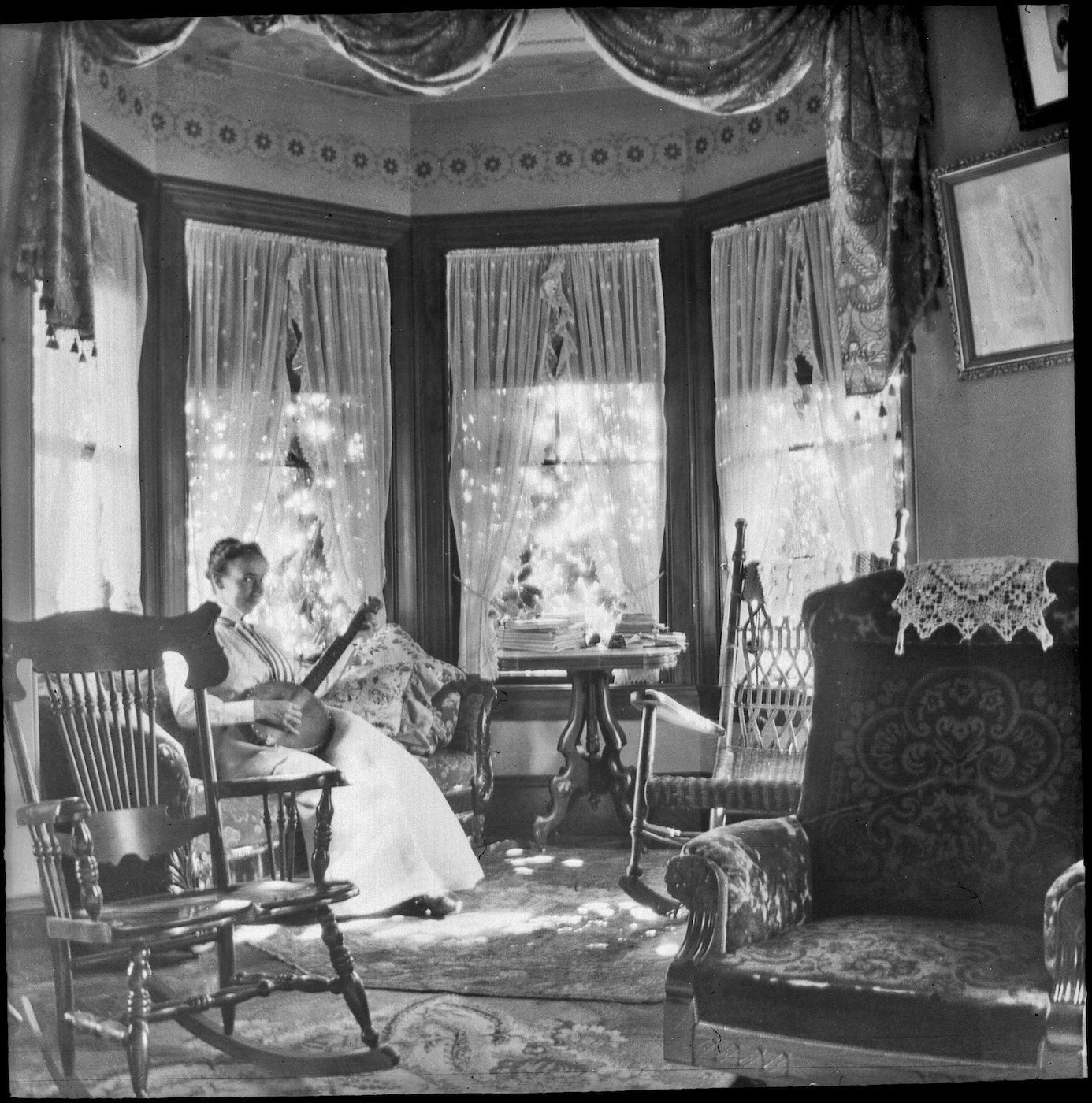 A black and white photograph of a woman seated in a rocking chair in a Victorian-era room.