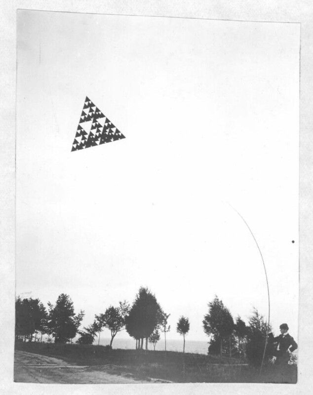 A black and white photograph of a large, triangular kite soaring in the sky above a line of trees and a person holding a long pole.
