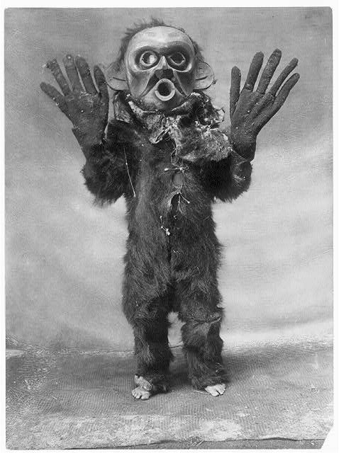 A full-body costume covered in dark, shaggy fur, with a pale mask featuring large eye sockets and an open mouth. The figure is standing with arms outstretched.