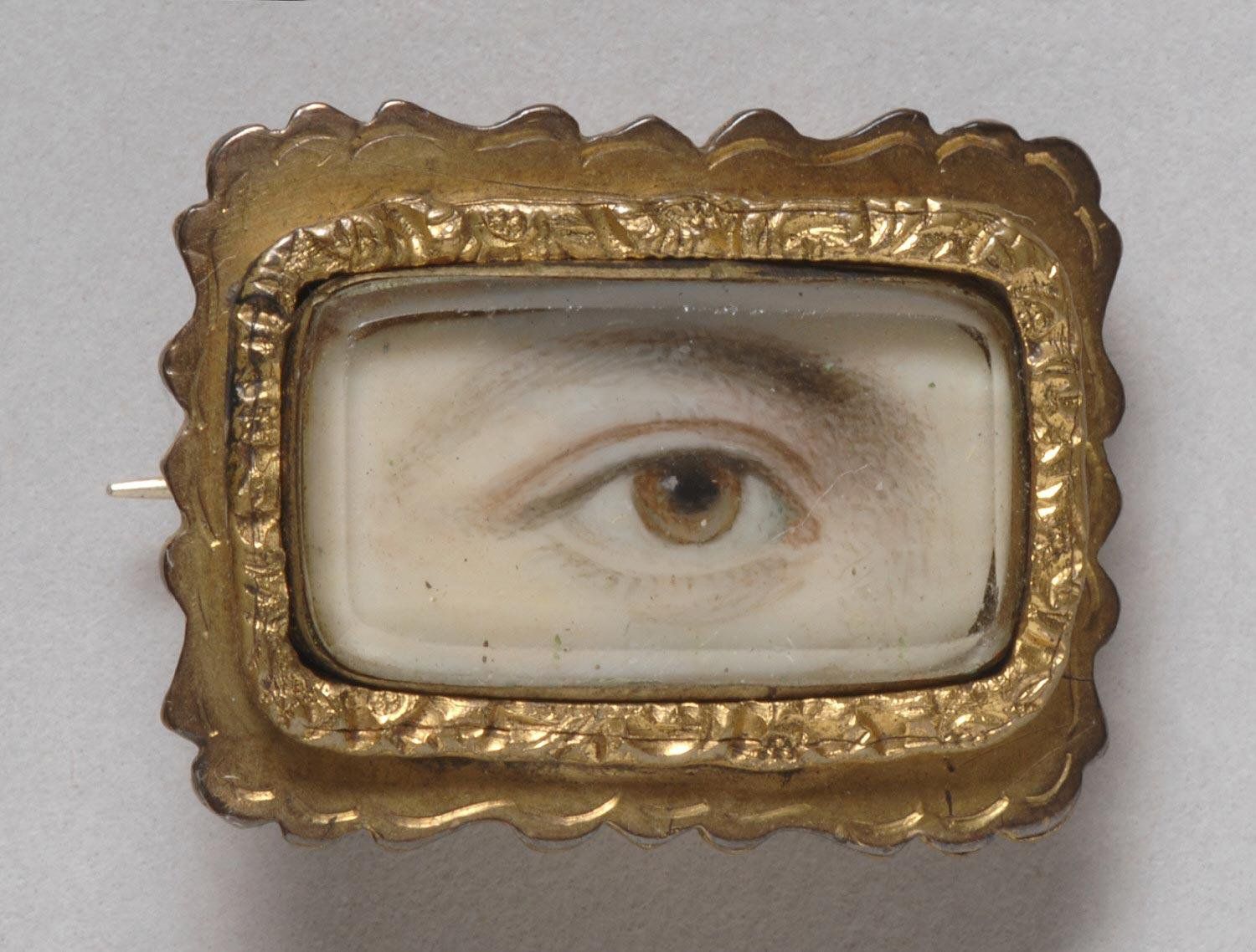 Close-up of a vintage brooch with a painted eye on ivory, set in a decorative gold frame.