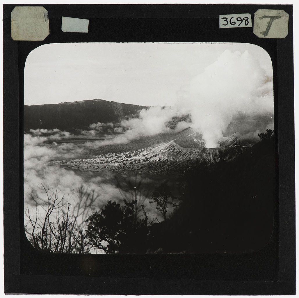 Black and white photograph of a volcano erupting, with a city at its base and a mountain range in the background.