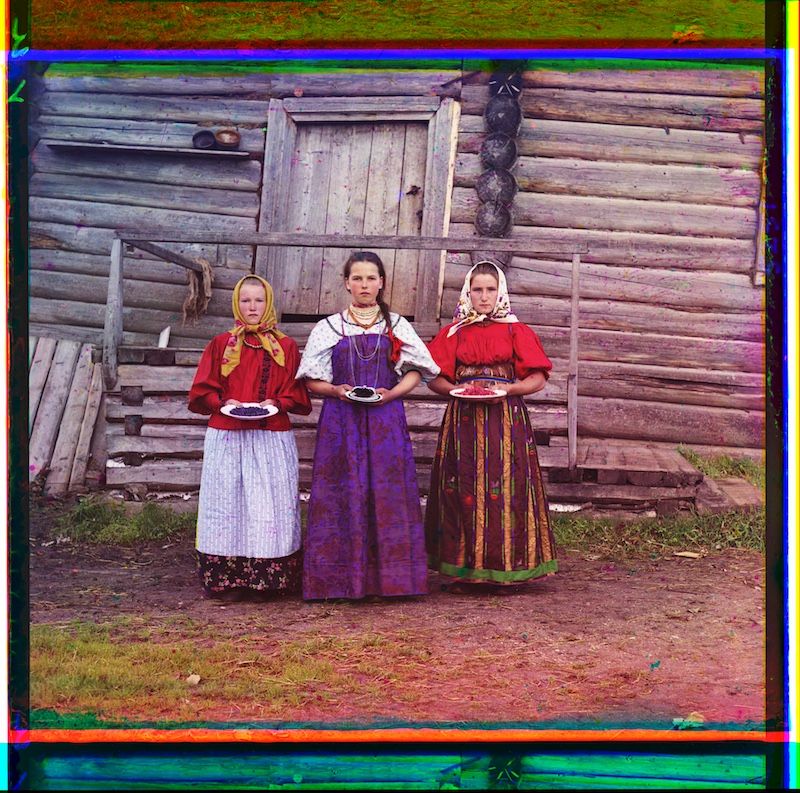 Three women in traditional folk costumes stand in front of a rustic wooden building, each holding a dish.