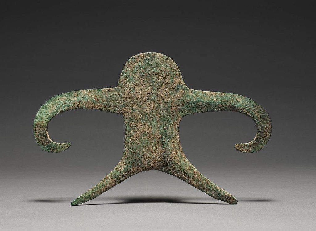 A bronze artifact shaped like a stylized human figure with arms extended horizontally and pointed feet, exhibiting a heavily patinated surface with shades of green, brown, and blue.