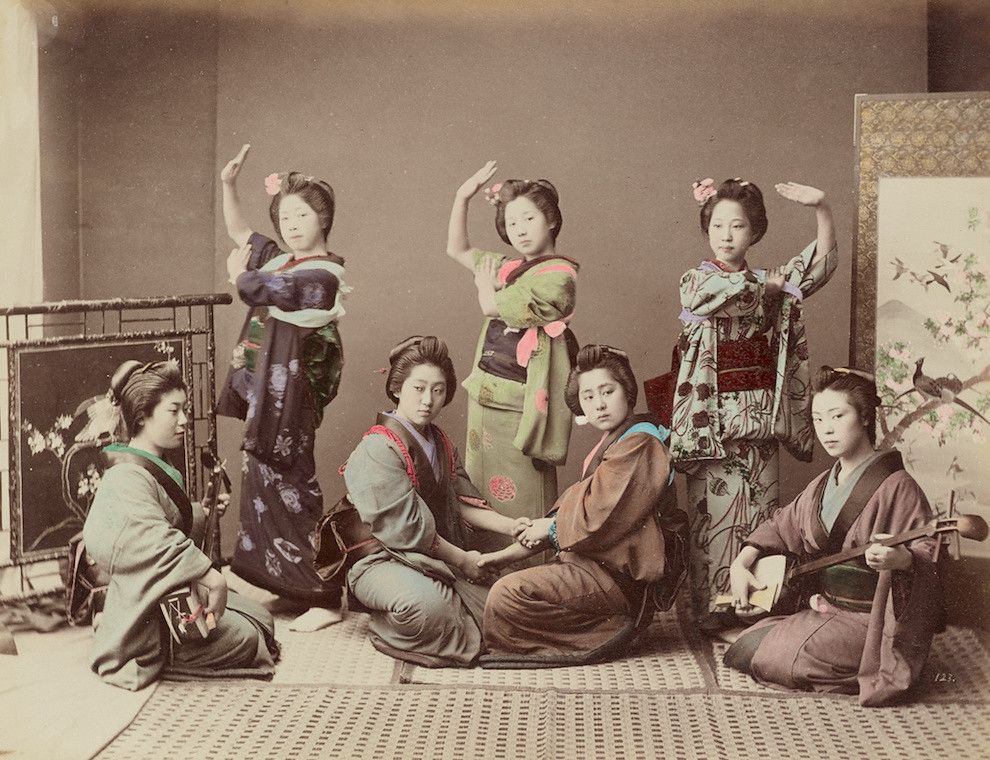 A vintage photograph of a group of Japanese women in traditional kimonos, posed in a studio setting with decorative screens and paintings.