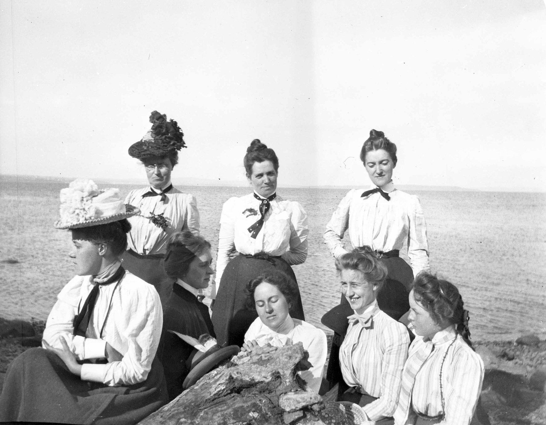 A vintage black and white photograph of seven women in Edwardian attire posing outdoors on a rocky outcrop overlooking water.