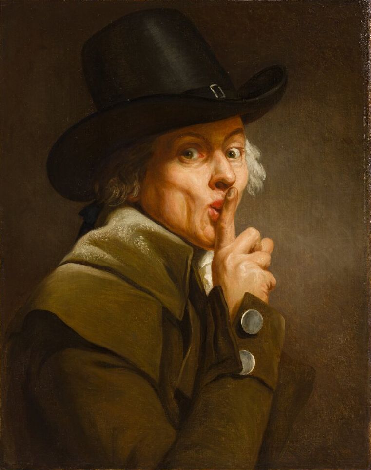 Portrait of a man in a black hat making a shushing gesture.