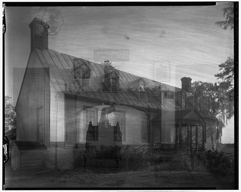 A black and white photograph of a large, stately house with a double exposure effect, revealing faint figures of people standing in front of the building.