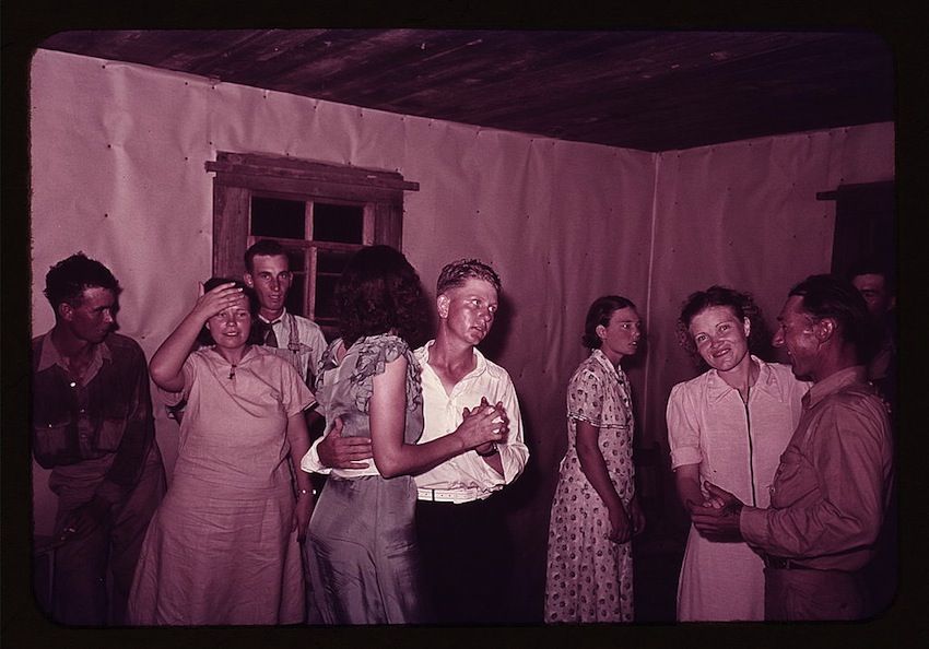 A vintage photograph capturing a lively scene inside a rustic room. The image is bathed in a distinctive pinkish hue, likely due to the film or lighting conditions of the time. The focus of the image is a group of people engaged in a lively dance.
