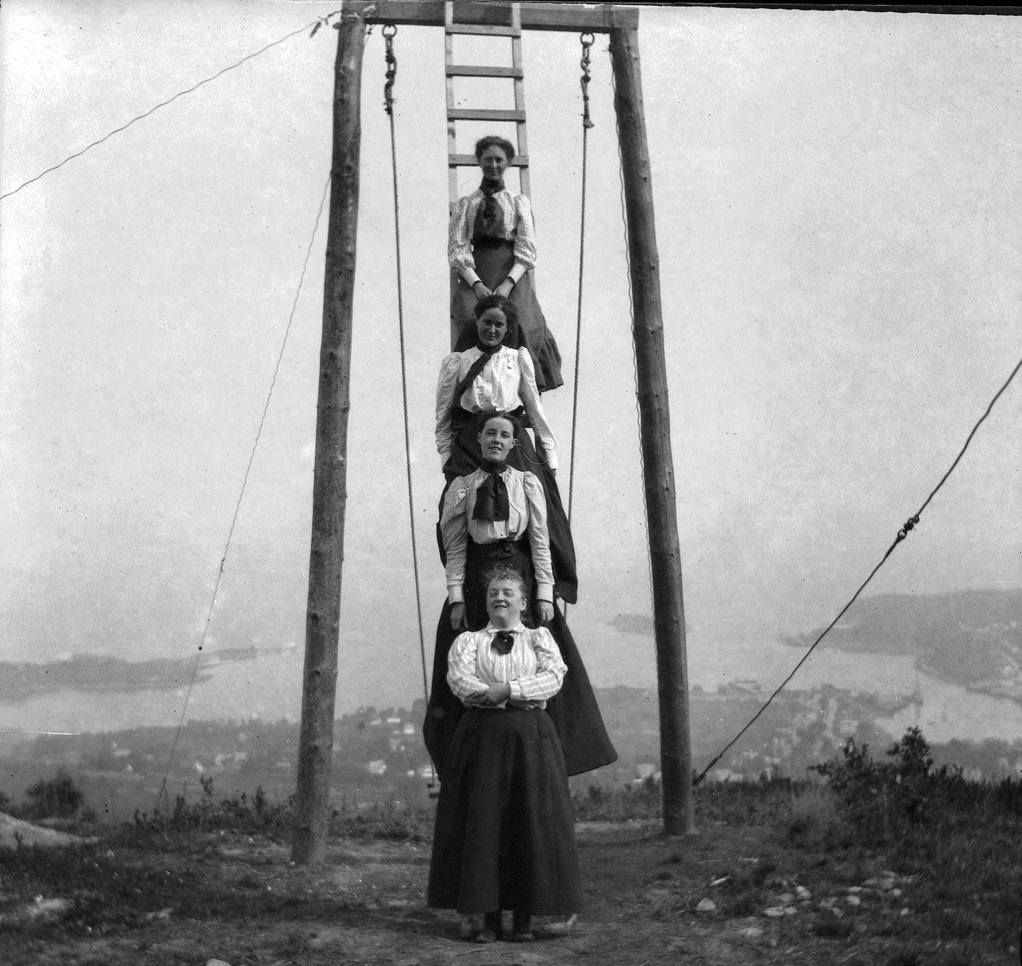 A vintage black and white photograph of four women stacked on top of each other on a wooden ladder-like structure on a hilltop.