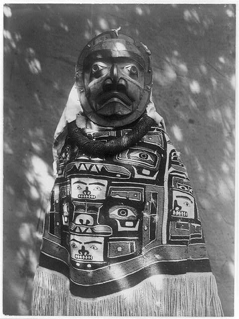 A black and white photograph of a person wearing a highly decorated mask and clothing, likely part of a ceremonial or cultural performance.