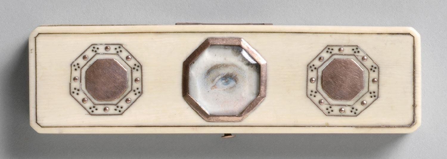 A rectangular ivory box with three octagonal elements. The central octagon contains a realistic depiction of a human eye, and the other two are made of copper-colored metal.