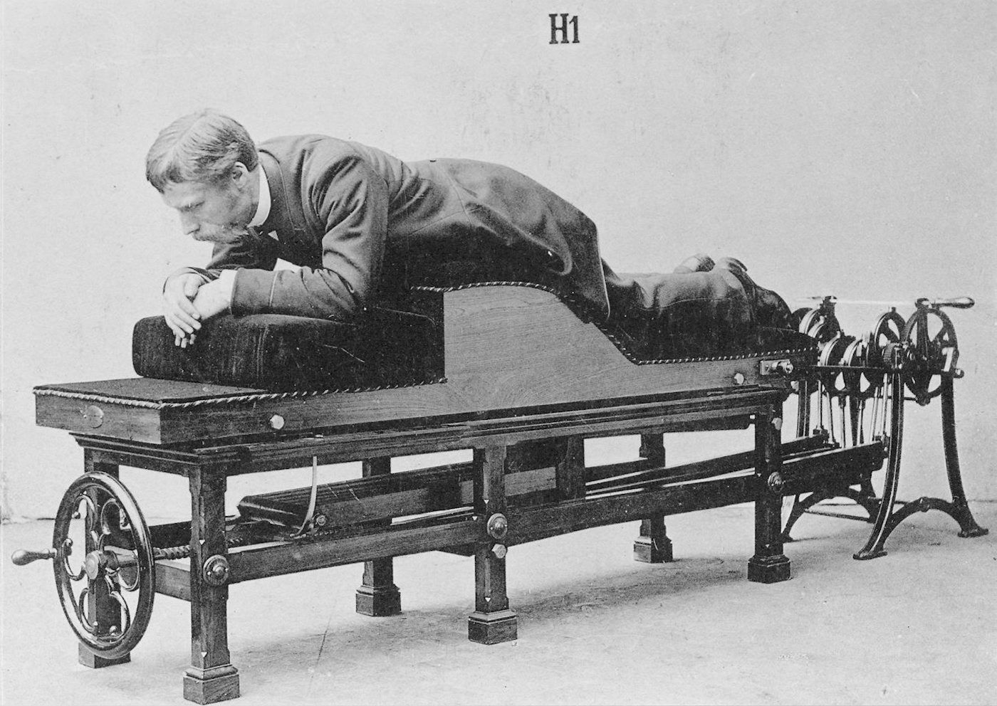 A vintage black and white photograph of a man lying head-down on a complex medical apparatus.