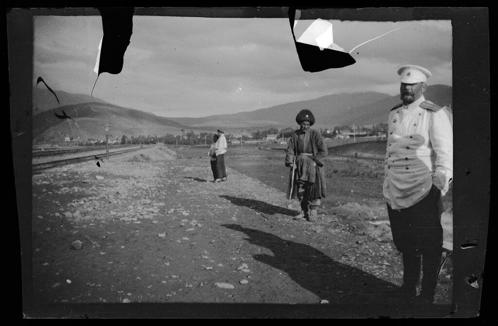 A black and white photograph of a landscape with several people in it. The photograph is damaged, with large tears and missing pieces along the top edge. The foreground features a rough, rocky field. A man in a military uniform stands prominently on the right, facing slightly away from the camera. He is wearing a peaked cap and a long coat, and appears to be an officer. His shadow is cast long on the ground. Further into the field, there are two other figures. One is a man wearing a traditional hat and a long coat, and the other is a person in a long dress or coat. They are walking away from the camera. The background consists of rolling hills and mountains, with a town or settlement visible in the distance. A railroad track runs along the base of the hills. The sky is bright and clear.