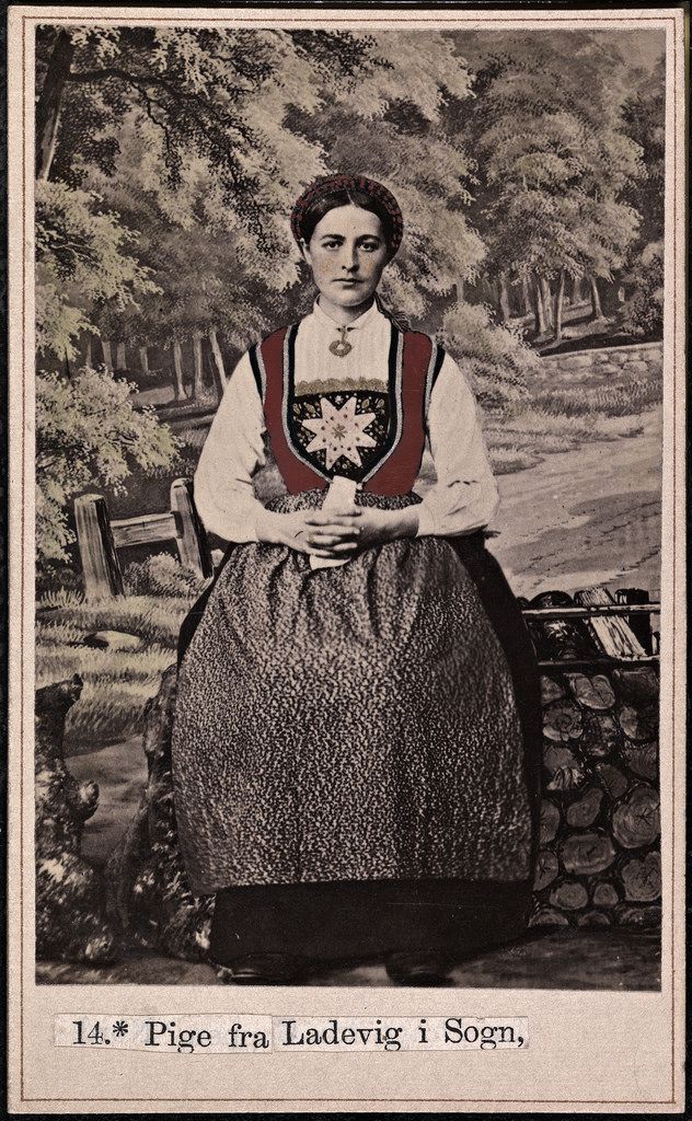 Vintage photograph of a young woman in traditional Norwegian folk costume standing in a grassy area with trees and a building in the background.
