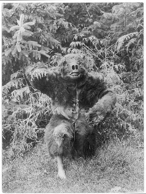 A vintage black and white photograph of a person in a full-body animal costume with a pig-like snout, posing in a grassy area with evergreen trees in the background.
