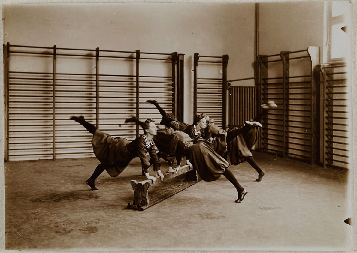 A sepia-toned photograph of a group of girls performing a leg lift exercise on a wooden bench in a gymnasium.