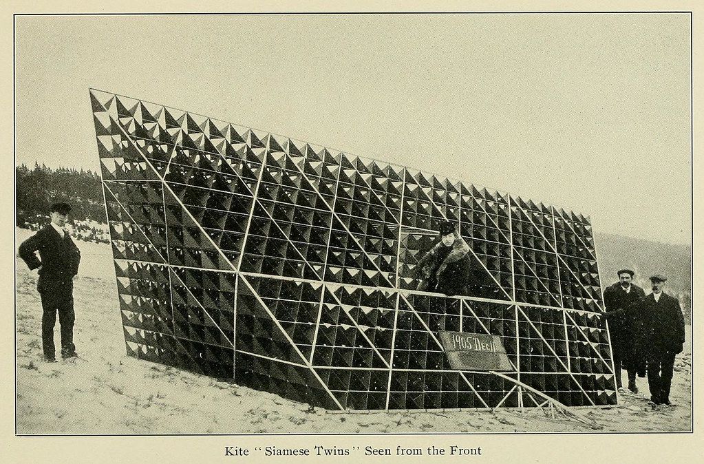 Vintage photograph of a large, geometrically complex kite titled "Siamese Twins" with a person inside, surrounded by three men in period clothing.