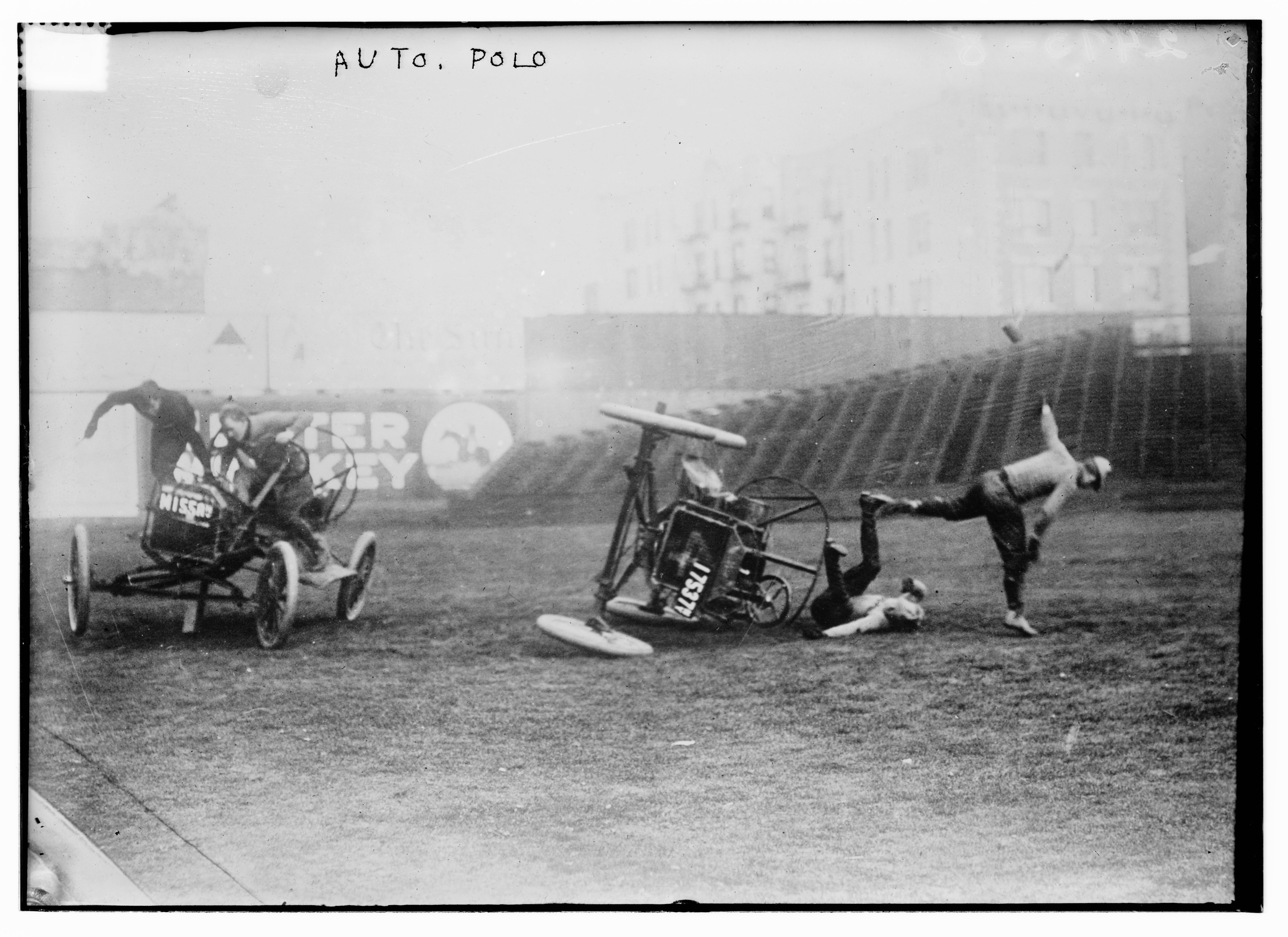 A grainy black and white photograph of an auto polo match. Two open-wheel cars are on a grassy field, one overturned with a man falling out. Another man stands nearby. The scene is labeled 'Auto Polo'.