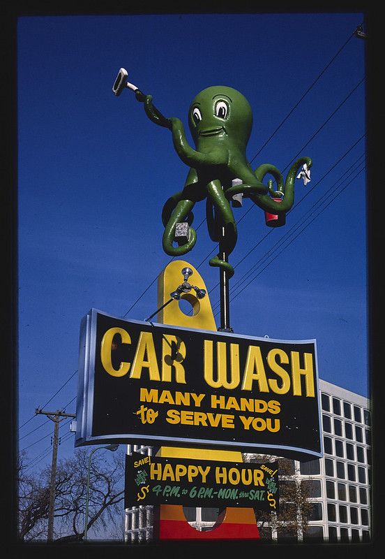 A vintage car wash sign featuring a green octopus with multiple arms holding car washing tools, set against a bright blue sky with a white building and bare trees in the background.