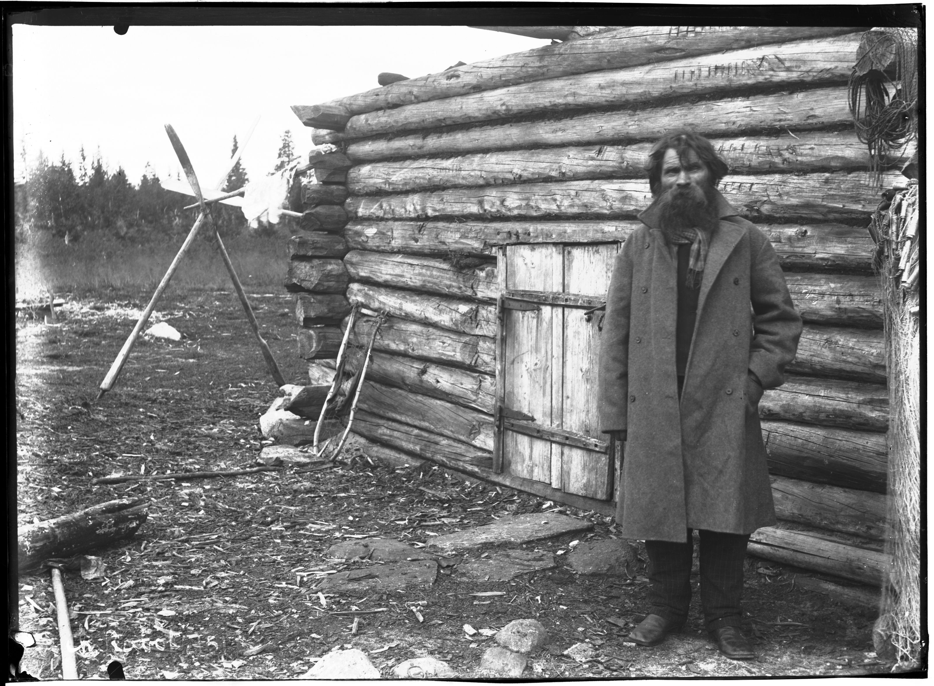 A black and white photograph of a man with a long beard standing in front of a log cabin with a wooden door. A primitive wind turbine stands to the left of the cabin. The ground is rocky and covered in debris, with trees in the background.