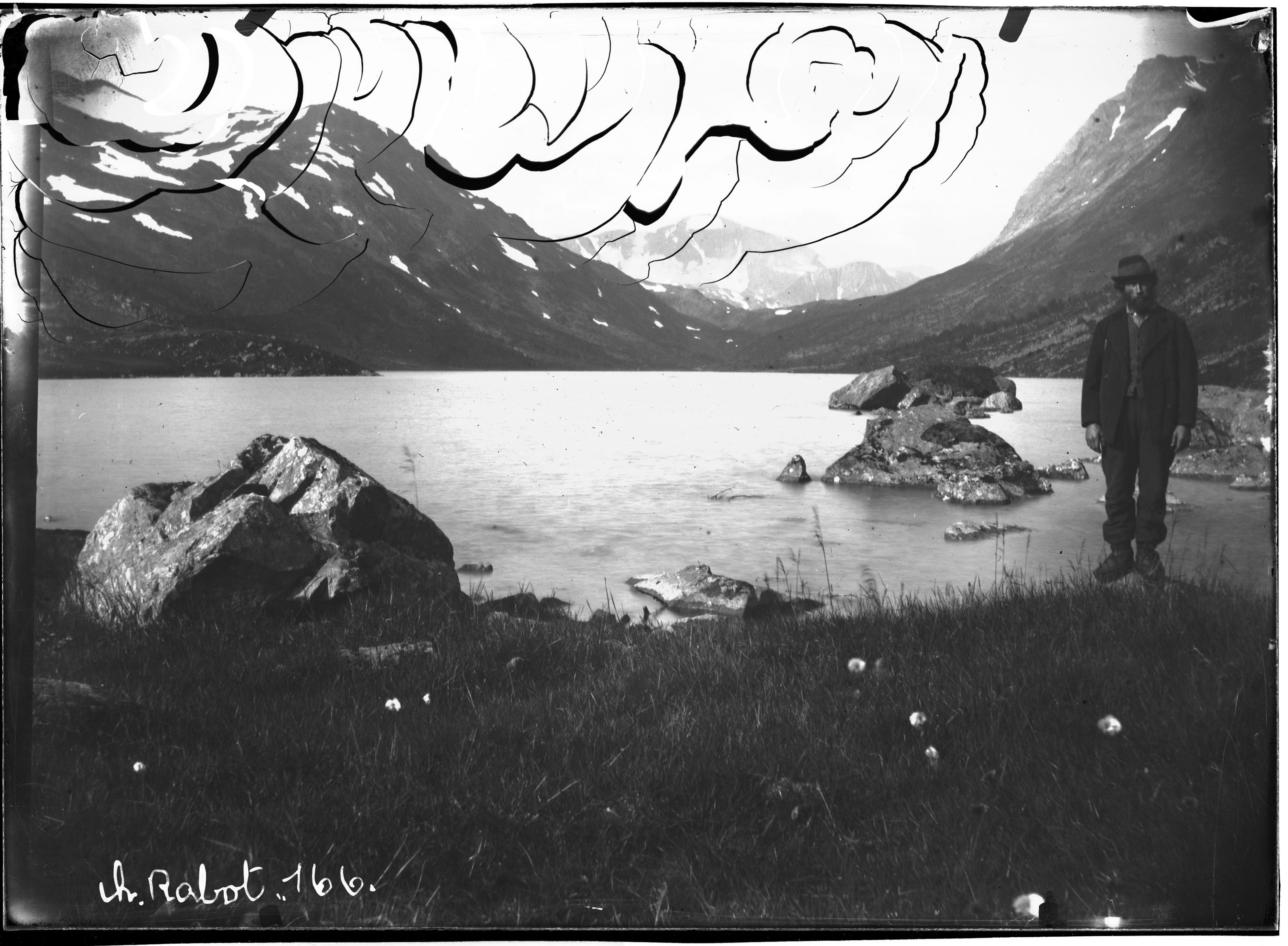 A vintage black and white photograph of a person standing by a lake surrounded by mountains.