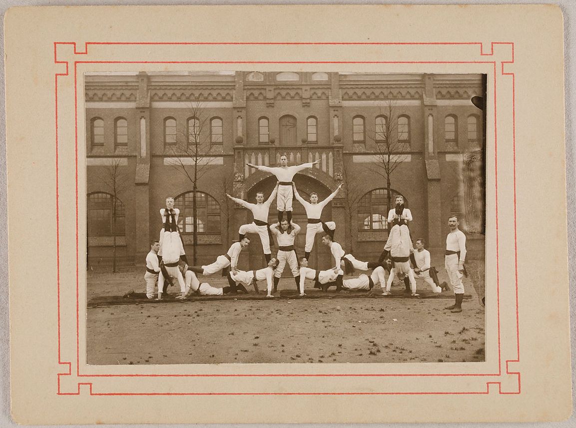 Vintage photograph of a group of young men forming a human pyramid in front of a brick building.