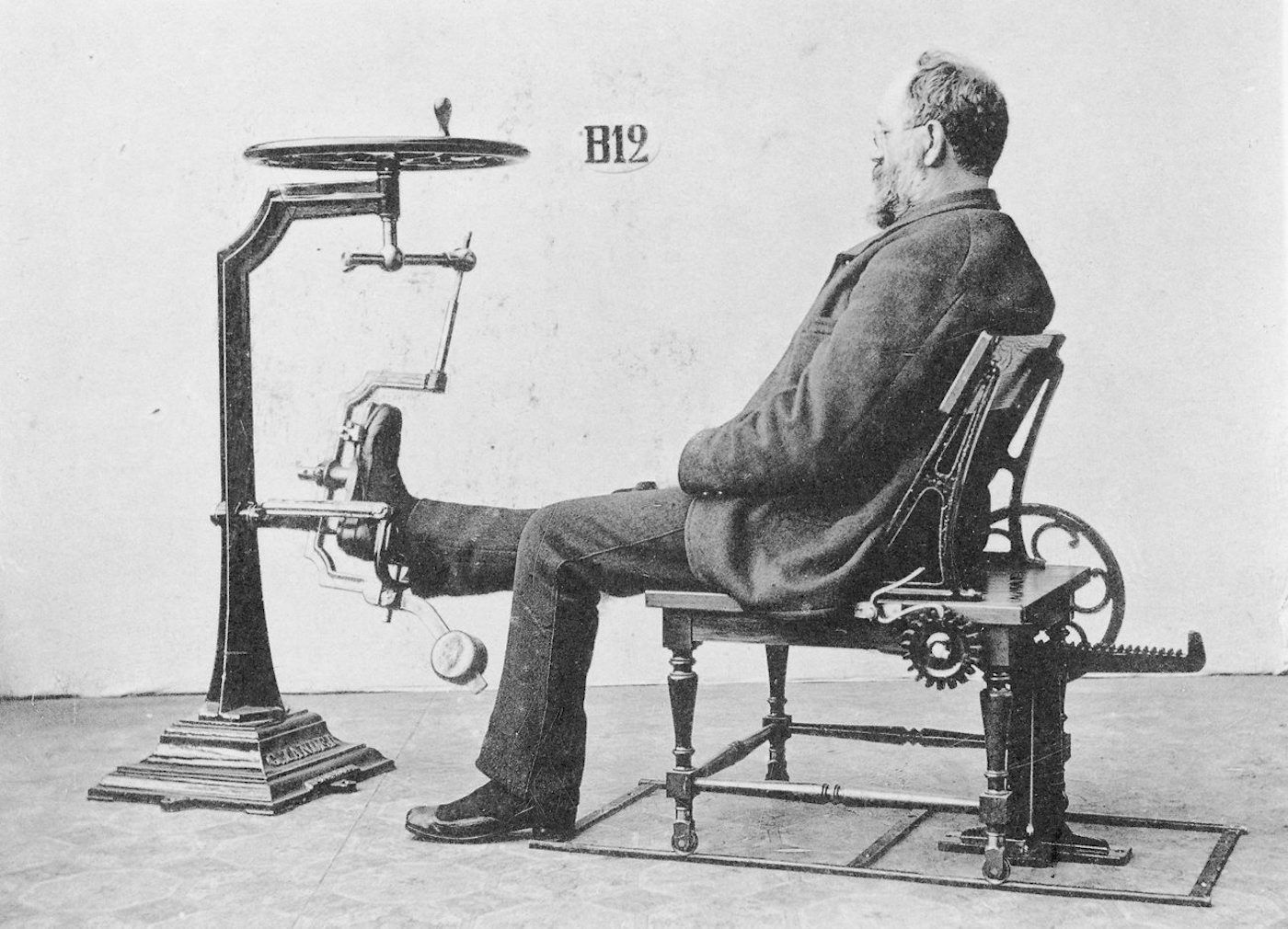 A black and white photograph of a man seated in a complex mechanical chair with a device attached to his leg.