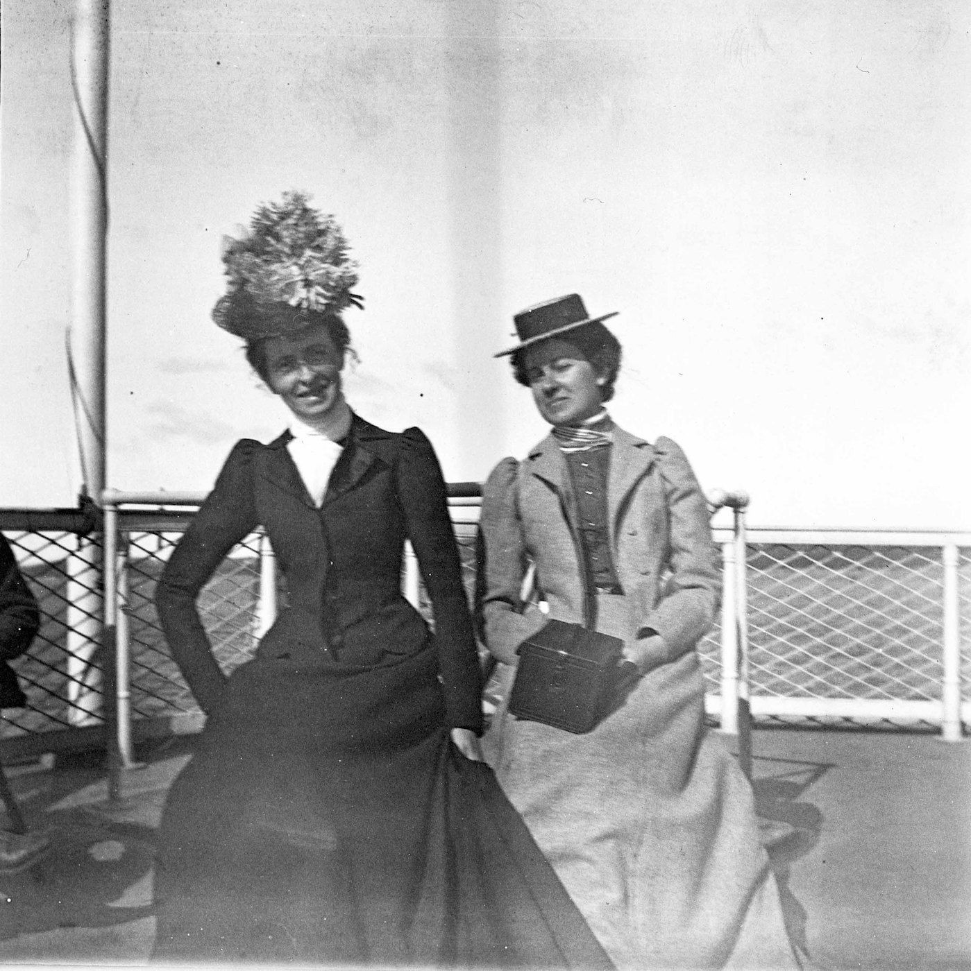 Two women in vintage clothing standing outdoors with a wire railing behind them.