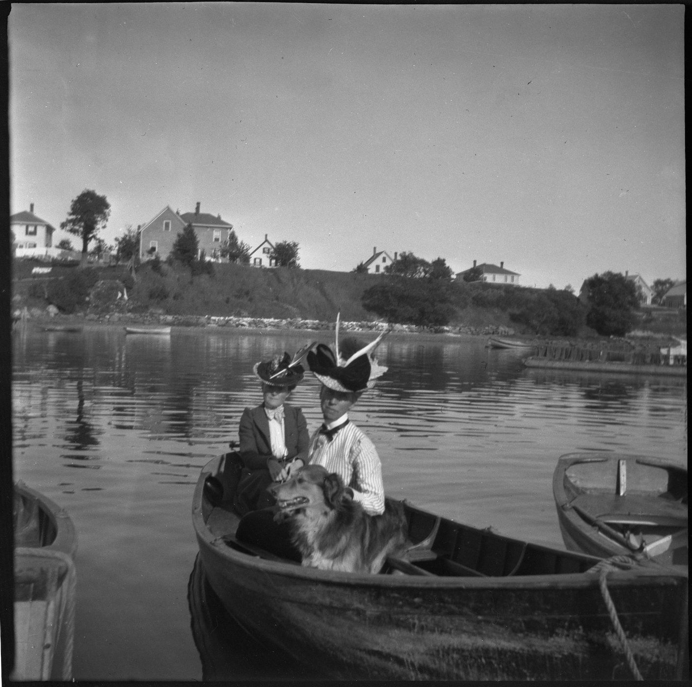 Black and white photograph of two people and a dog in a boat on a lake, with houses and trees in the background.
