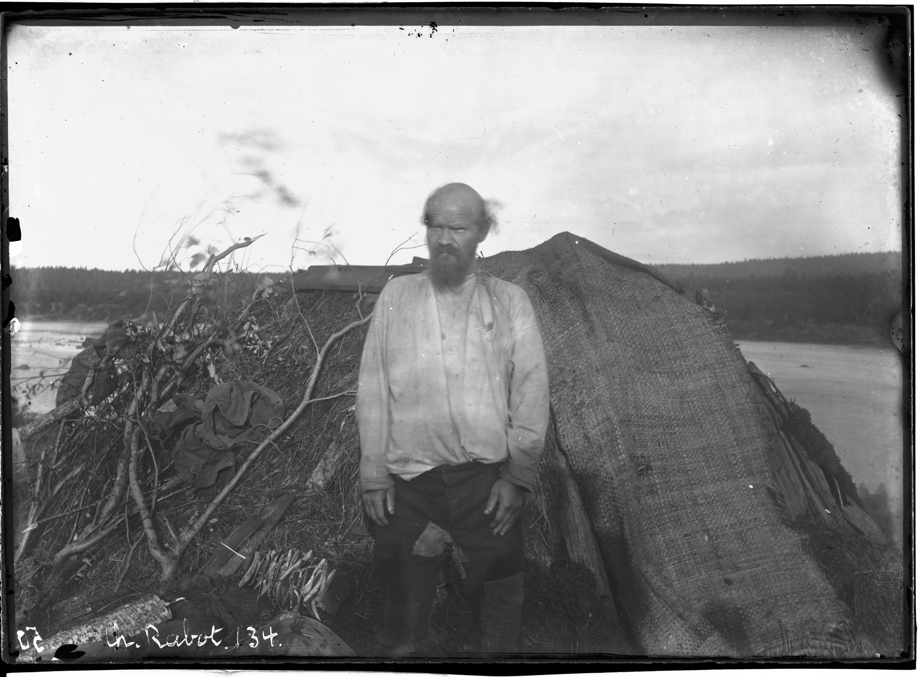A vintage black and white photograph of an older man with a long beard standing in front of a thatched-roof hut in a forest.