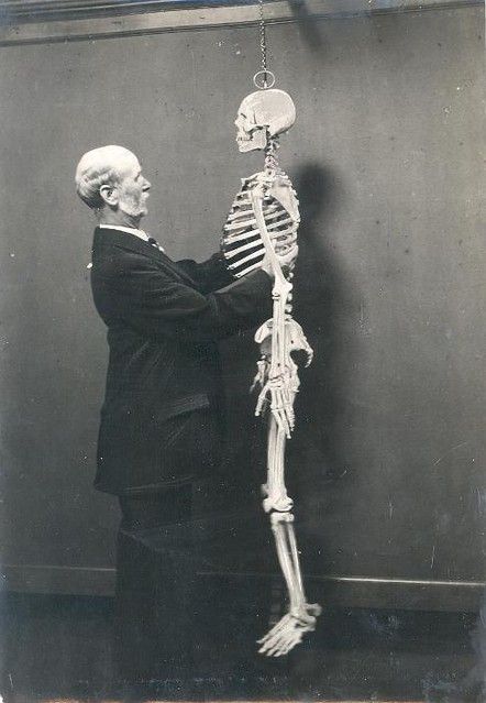 A vintage black and white photograph of a man standing next to a full human skeleton.